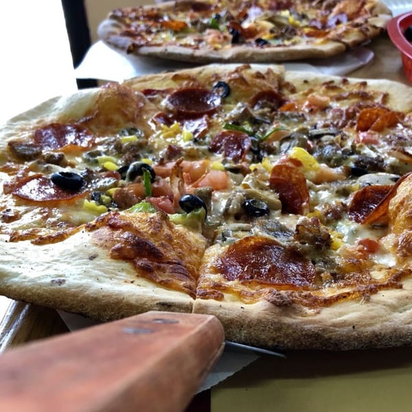 Two freshly baked pizzas, one in focus with pepperoni, olives, and other toppings, on a table.
